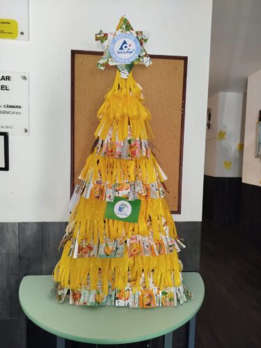 A árvore evidencia o envolvimento ativo dos alunos na construção de um elemento decorativo sustentável. A escolha da cor amarela confere destaque visual à estrutura e valoriza a identidade das embalagens utilizadas. promovendo a reutilização de materiais