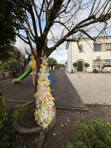 Árvore da Natal no recinto escolar exterior, com o parque infantil e a escola ao fundo.