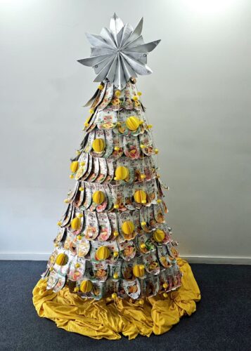 Árvore de Natal feita com uma estrutura de madeira triangular, rede de galinheiro, pacotes de sumo Compal, trapilho, tampas de plástico, restos de papel amarelo, fio amarelo e malha amarela.