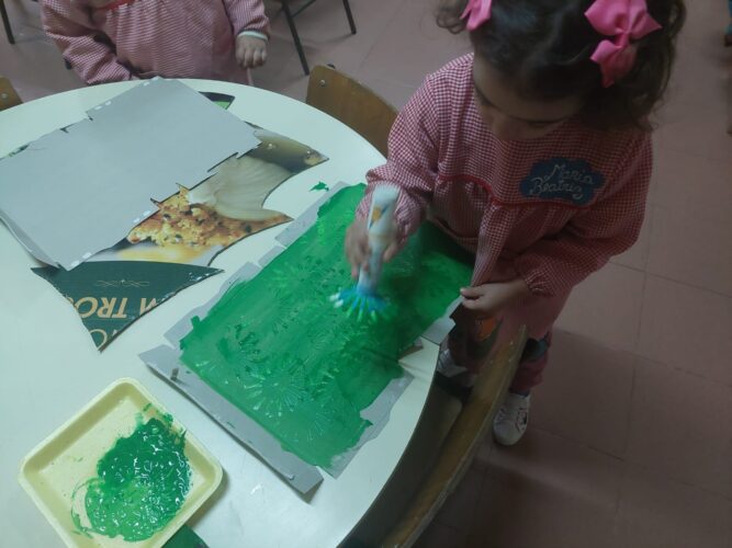 Criança a pintar uma caixa de cereais, com pincel e guache verde claro e verde escuro, para elaboração da relva para o painel.