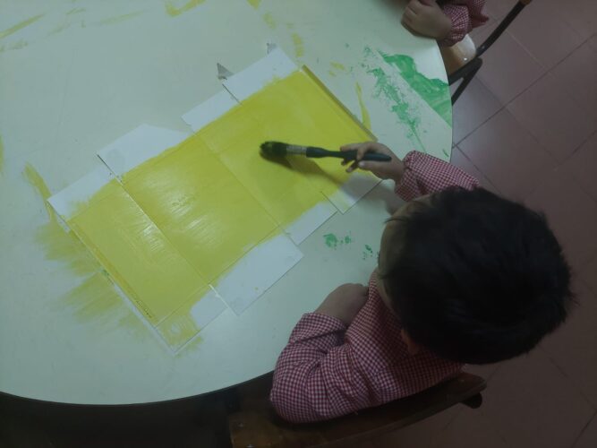 Criança a pintar uma caixa de cereais, com pincel e guache amarelo, para a elaboração dos elementos decorativos e título do painel.