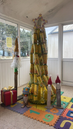 Árvore de Natal construída com embalagens Compal, pintada de amarelo e decorada com estrela no topo, destacando o logótipo “Compal – Em Família”, trazendo alegria, cor e espírito natalício ao espaço da escola.