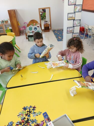 Processo de construção da Árvore de Natal. Crianças a cortar embalagens Compal.<br/>As crianças realizaram cortes e transformações com apoio tendo desenvolvido a motricidade fina, a autonomia e a consciência da reutilização de resíduos como recurso criativo.