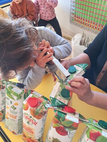 Criança a participar ativamente na construção da Árvore de Natal Amarela. <br/>Agrafa as embalagens para que fiquem ligadas, com apoio do adulto. Permite compreender como a reutilização pode fornecer nova função estrutural ao material e promover a cooperação