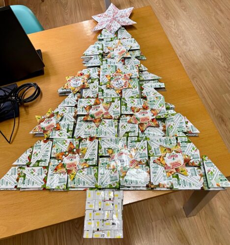 Árvore de Natal feita com cartão grosso e decorada com pacotes de 200 mL da Compal. As estrelas, destacando tons de amarelo, decoram a árvore, sendo a principal marcada com as palavras Tetra Pak e Compal. O tronco foi formado a partir das riscas amarelas
