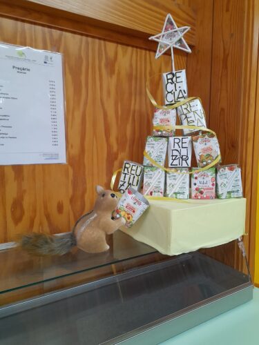 Um esquilo interage com a árvore e acrescenta um toque apelativo à exposição. À esquerda, vê-se um preçário do bar. O conjunto cria uma narrativa visual que cruza consumo, reutilização e consciência ambiental num espaço comum da escola – o bar dos alunos