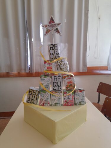Embalagens Tetra Pak da Compal, dispostas em pirâmide sobre uma base amarela e um fita também amarela serpenteia à volta da estrutura. As palavras ambientais: “REDUZIR”, “REUTILIZAR”, “RECICLAR” e “REPENSAR” e uma estrela feita com palhinhas recicláveis.